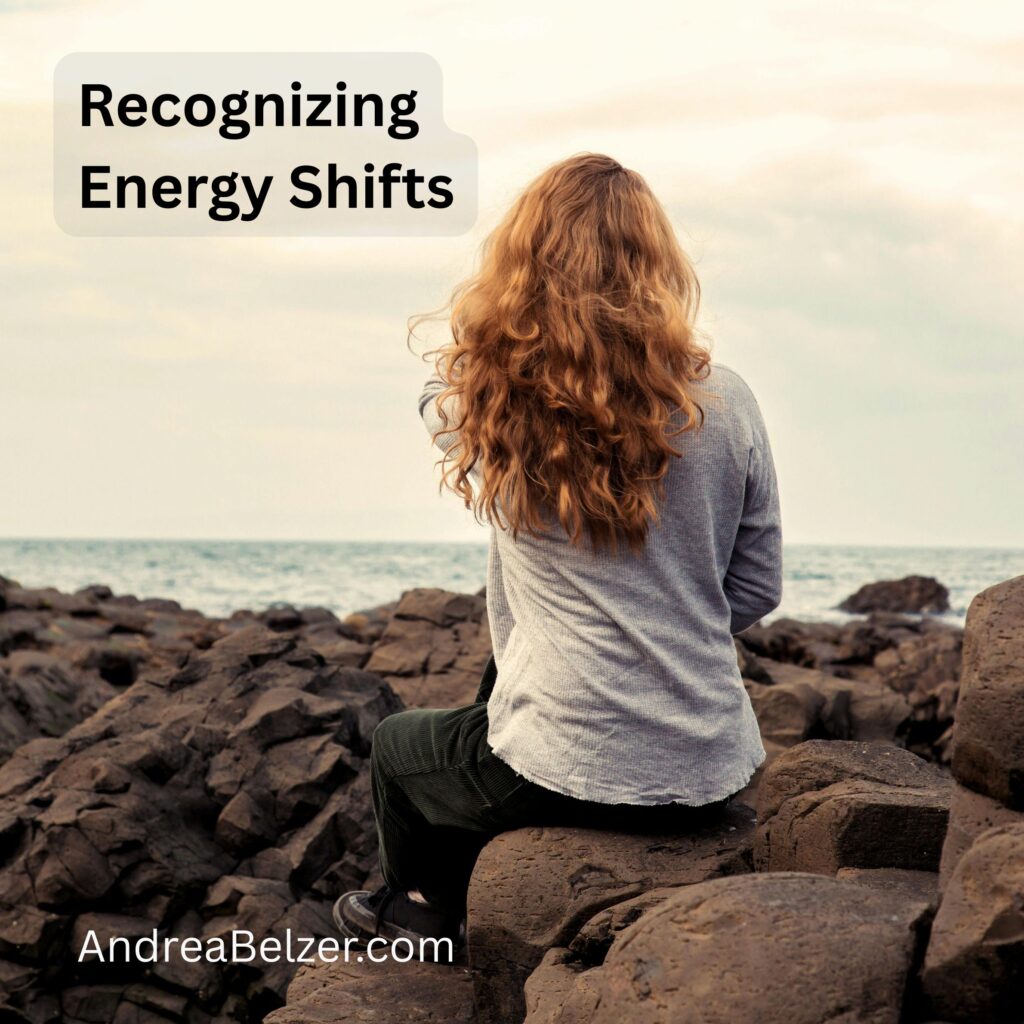 Woman sitting on rocks by the ocean with back to camera. Text overlay says - "Recognizing energy shifts" and "andreabelzer.com"