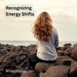 Woman sitting on rocks by the ocean with back to camera. Text overlay says - "Recognizing energy shifts" and "andreabelzer.com"